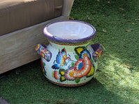 A colorful Talavera ceramic flower pot with a floral pattern featuring hummingbirds and lavender trim, displayed on a green surface.