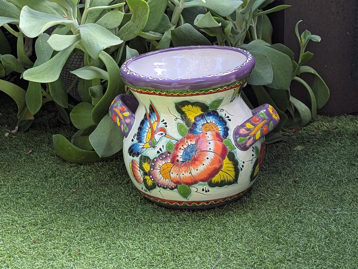 A colorful Talavera ceramic flower pot with a floral pattern featuring hummingbirds and lavender trim, displayed on a green surface.