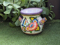A colorful Talavera ceramic flower pot with a floral pattern featuring hummingbirds and lavender trim, displayed on a green surface.
