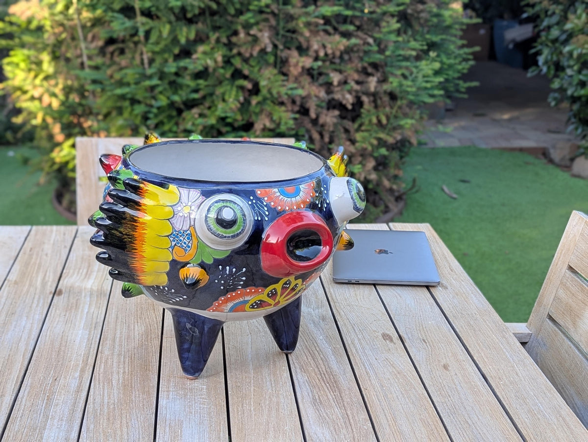 A hand-painted Talavera ceramic planter in the shape of a blowfish with a colorful and decorative design, placed on a wooden surface outdoors.