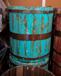 A distressed turquoise wooden barrel with dark metal bands, shown in a storage setting next to other barrels.