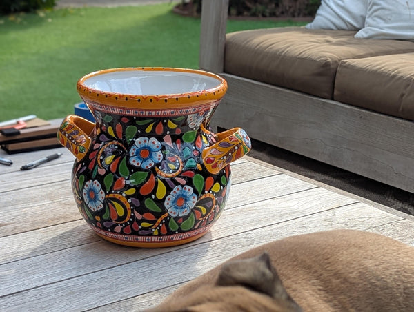 A colorful, round Talavera ceramic flower pot with blue trim and handles, displayed on a wooden surface with a dog lying in front of it.