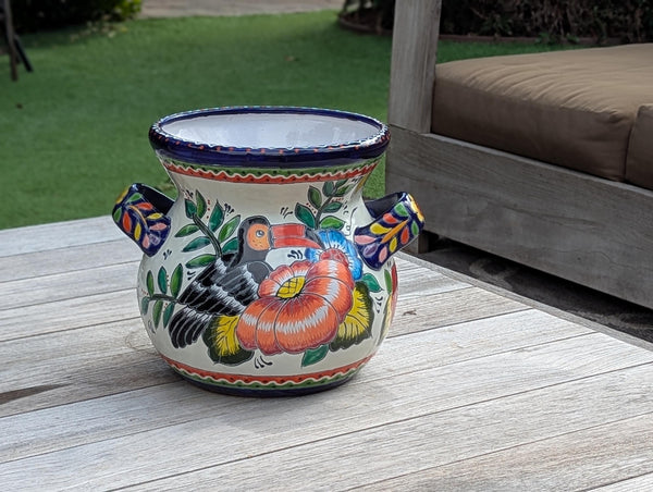 A round ceramic planter with handles featuring a colorful floral and toucan design, placed on outdoor furniture.